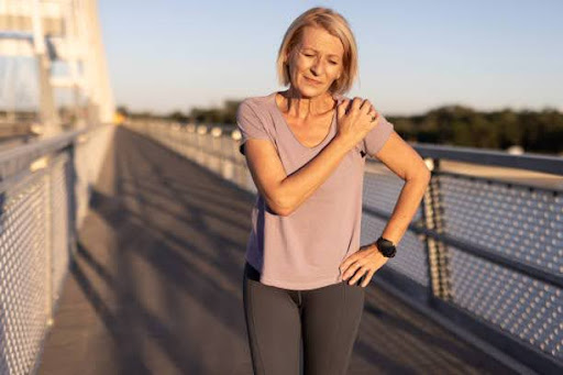 A senior woman dressed in workout attire holds her shoulder in pain while standing on a bridge during golden hour, capturing a moment of discomfort and strain during exercise.